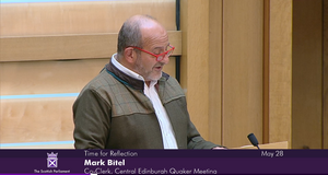 Mark speaking to Parliament Mark Bitel, Co-Clerk of Central Edinburgh Quaker Meeting, addresses Scottish Parliament. Mark is wearing a white shirt, green jacket and red glasses.